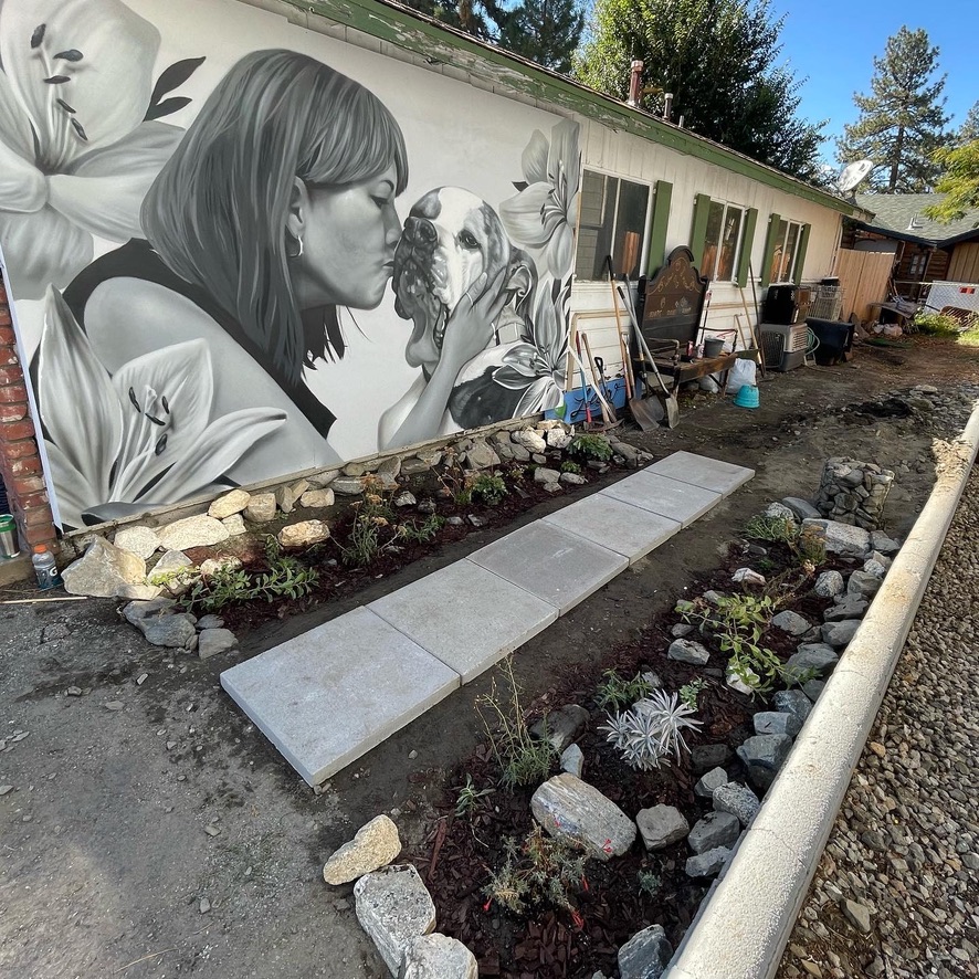 A wall covered in a detailed and airbrushed style of Lesa Dawn Smith giving her pitbull a big smooch, surrounded by beautiful flowers. The garden is in the area in front of it, with a pathway going through the center of it